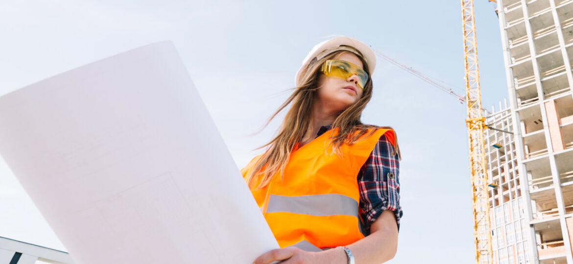 Construction worker reviewing blueprints at site wearing safety gear.